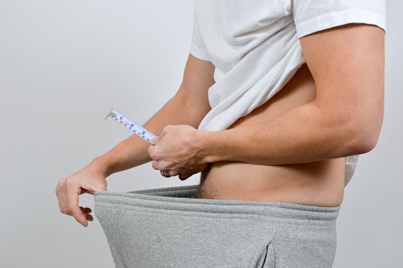 A man holding a measuring tape above his crotch and stretching the waistband on his jogging bottoms to imply that he is measuring his penis size