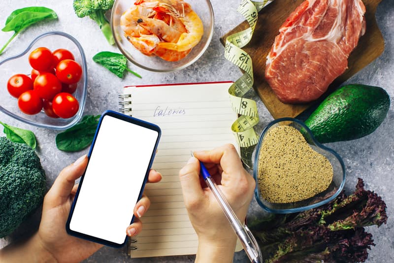 A hand writing on a notepad to track progress, with another hand holding a mobile phone. Surrounded by healthy foods.