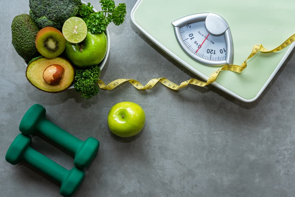 A pair of weights, an apple and a bowl of healthy fruits and vegetables next to a weighing scale