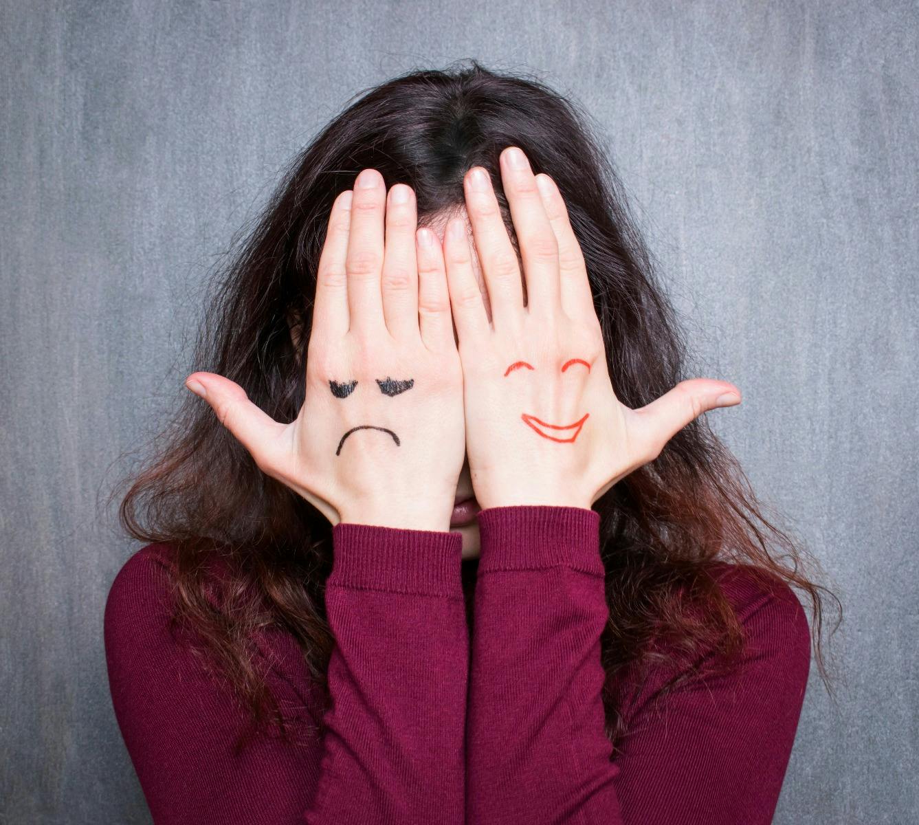 Woman covering her face with her hands. One hand has a happy face and one hand has an angry face.