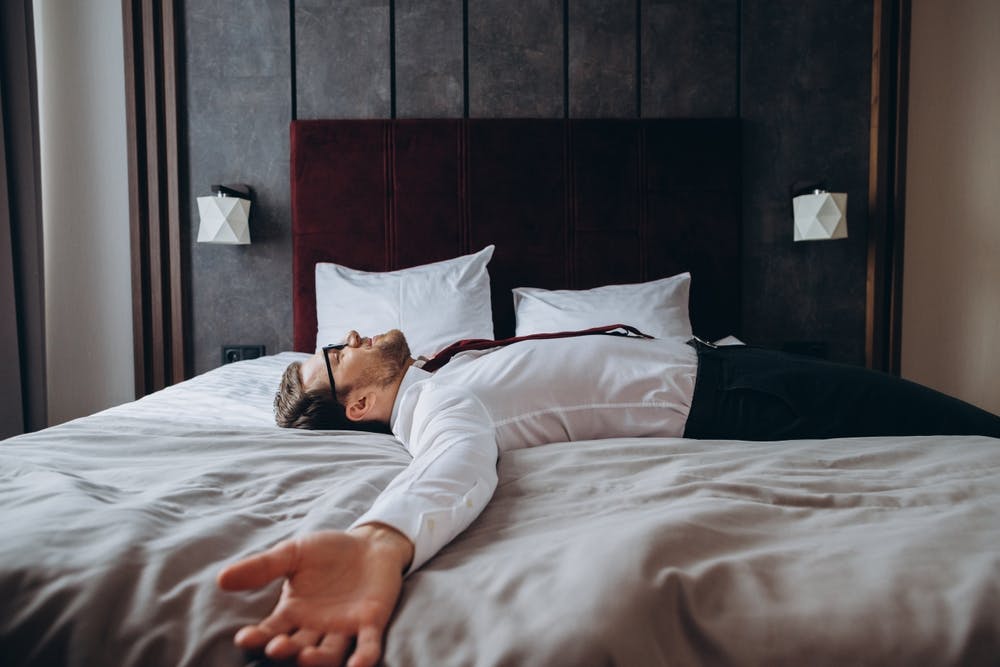 Man lying on a bed with a shirt and tie on, experiencing the side effects of jet lag
