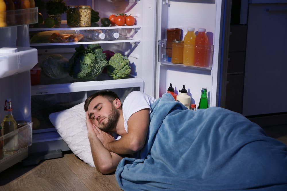 Man sleeping next to an open fridge with lots of food inside it