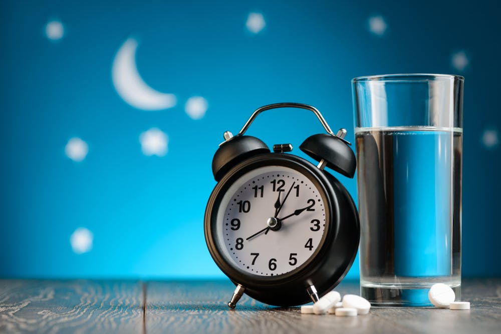 Alarm clock showing the time 00:10, next to a glass of water and in front of a background containing the moon and stars at night time